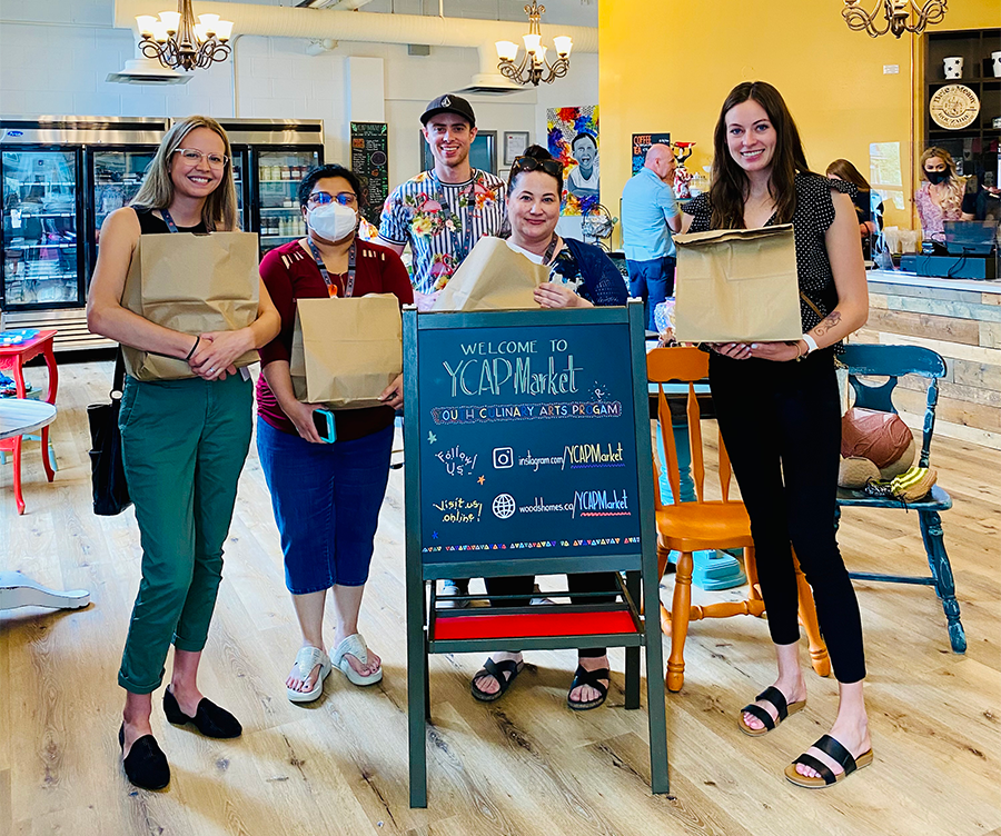 img des: A group of 5 people standing in front of a sign for the market, holding shopping bags.