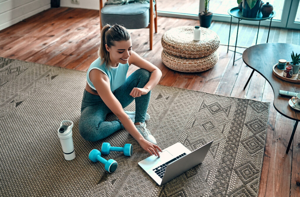 A woman sitting in her living room with dumbbells, a water bottle, and laptop, ready to do a workout class to help cope with crippling anxiety.